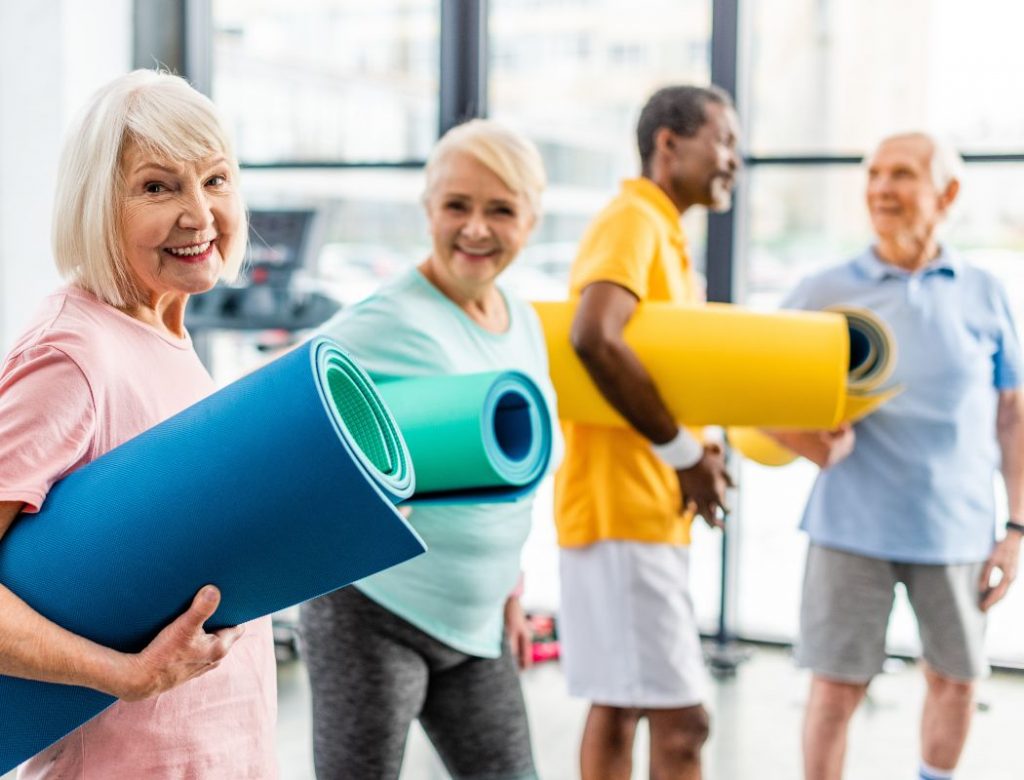 selective focus of senior sportswoman holding fitness mat and her friends standing behind at gym