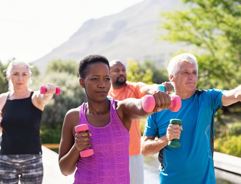 Happy senior and mature couples exercising with dumbbells. Healthy multiethnic people exercising using dumbbells outdoor. African couple and senior friends in sportswear stretching arms holding colorful dumbbells at park.